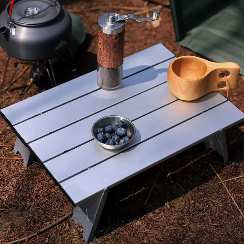 Compact aluminum folding camping table with coffee grinder, wooden cup, and blueberries. Ideal for outdoor recreation and hiking.