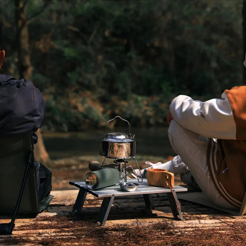 Campers sitting beside Ultralight Folding Camping Table with cooking gear. Compact aluminum desk perfect for outdoor adventures.